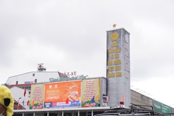 A large building facade with multiple signs, including an advertisement with bright orange colors and Vietnamese text. The building features a vertical structure with a golden star at the top and more text in yellow on a gray background. Several flags are situated near the building, and a vehicle appears in the foreground.