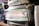 A close-up of a stainless steel espresso machine features the brand name 'E61' prominently on the front panel. The machine is situated on a wooden counter and includes a hopper filled with coffee beans. Stacked disposable coffee cups are visible to the right.