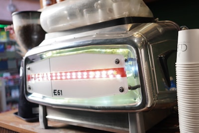 A close-up of a stainless steel espresso machine features the brand name 'E61' prominently on the front panel. The machine is situated on a wooden counter and includes a hopper filled with coffee beans. Stacked disposable coffee cups are visible to the right.