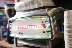 A close-up of a stainless steel espresso machine features the brand name 'E61' prominently on the front panel. The machine is situated on a wooden counter and includes a hopper filled with coffee beans. Stacked disposable coffee cups are visible to the right.