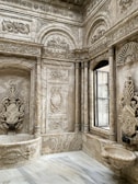 Elaborate interior space featuring intricately carved stone walls with floral and geometric patterns. The design includes arches, columns, and a large window that lets in natural light. The overall architecture displays a luxurious and ornate style.