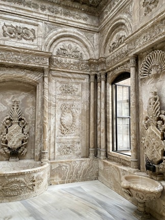 Elaborate interior space featuring intricately carved stone walls with floral and geometric patterns. The design includes arches, columns, and a large window that lets in natural light. The overall architecture displays a luxurious and ornate style.