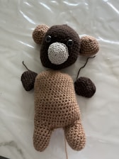 A handmade crocheted bear is lying on a smooth, white surface. It has a brown body, darker brown arms and head, lighter brown ears, and a cream-colored muzzle. The bear features black eyes and an expressionless face.
