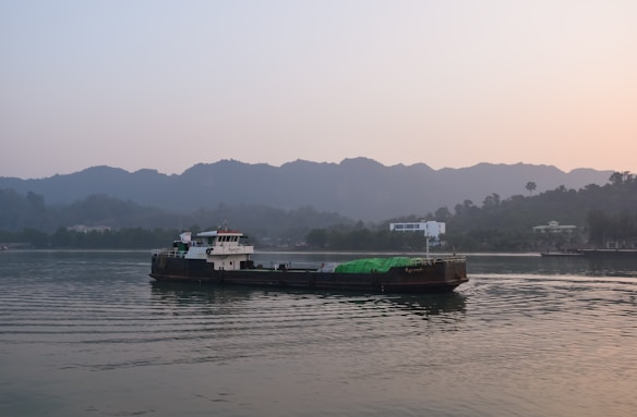 A large cargo ship sails on calm waters, with a green tarpaulin covering part of its deck. The background features rolling mountains partially shrouded in mist, creating a serene and tranquil scene. The sky is clear with soft light, suggesting either early morning or late afternoon.