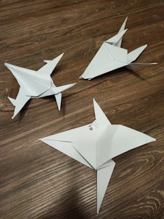 Three paper origami models resembling shurikens are placed on a wooden surface. The models are crafted from white paper, showcasing precise folds and pointed edges. A small mark or writing is visible on one of the models.