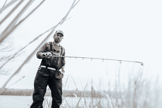 A person wearing outdoor gear and a cap is holding a fishing rod while standing near a body of water. The image has a soft, muted color palette, creating a serene atmosphere. Tall grasses are visible in the foreground, and the person looks focused on their activity.