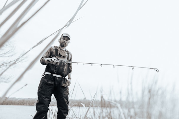 A person wearing outdoor gear and a cap is holding a fishing rod while standing near a body of water. The image has a soft, muted color palette, creating a serene atmosphere. Tall grasses are visible in the foreground, and the person looks focused on their activity.