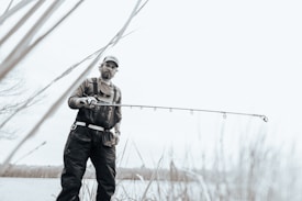 A person wearing outdoor gear and a cap is holding a fishing rod while standing near a body of water. The image has a soft, muted color palette, creating a serene atmosphere. Tall grasses are visible in the foreground, and the person looks focused on their activity.