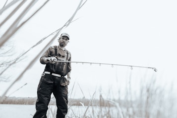 A person wearing outdoor gear and a cap is holding a fishing rod while standing near a body of water. The image has a soft, muted color palette, creating a serene atmosphere. Tall grasses are visible in the foreground, and the person looks focused on their activity.