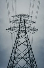 A towering metal electricity pylon surrounded by multiple power lines against a cloudy sky. The structure is symmetrical and prominently rises into the frame, showcasing its intricate crisscrossing beams and sturdy construction.