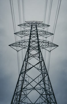A towering metal electricity pylon surrounded by multiple power lines against a cloudy sky. The structure is symmetrical and prominently rises into the frame, showcasing its intricate crisscrossing beams and sturdy construction.