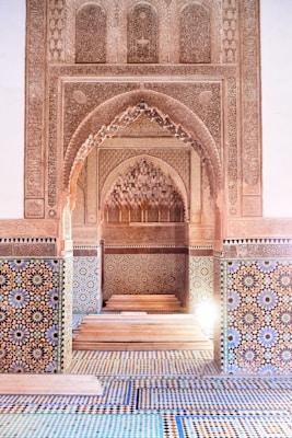 Intricate architecture featuring ornate arches and detailed tile work with geometric patterns. The walls are adorned with elaborate carvings, and the floor displays complex mosaic designs in blue, green, and earthy tones. A soft light enhances the sophisticated craftsmanship.
