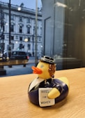 A rubber duck dressed in a historical outfit is perched on a wooden surface. It wears a black hat and a suit, holding a sign that reads 'Votes for Women.' A large building is visible through the window in the background, with a street scene including a car and some people walking by.
