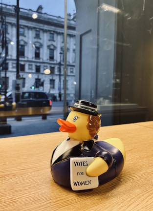 A rubber duck dressed in a historical outfit is perched on a wooden surface. It wears a black hat and a suit, holding a sign that reads 'Votes for Women.' A large building is visible through the window in the background, with a street scene including a car and some people walking by.