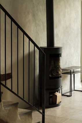 A modern interior space featuring a black metal staircase railing adjacent to a cylindrical black wood-burning stove. The stove stands against a smooth, light-colored wall, and a small bench is placed nearby.