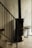 A modern interior space featuring a black metal staircase railing adjacent to a cylindrical black wood-burning stove. The stove stands against a smooth, light-colored wall, and a small bench is placed nearby.
