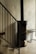A modern interior space featuring a black metal staircase railing adjacent to a cylindrical black wood-burning stove. The stove stands against a smooth, light-colored wall, and a small bench is placed nearby.
