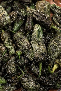 A collection of blistered green peppers covered in coarse salt, piled together. The peppers have a shiny surface, indicating they have been cooked and perhaps oiled.