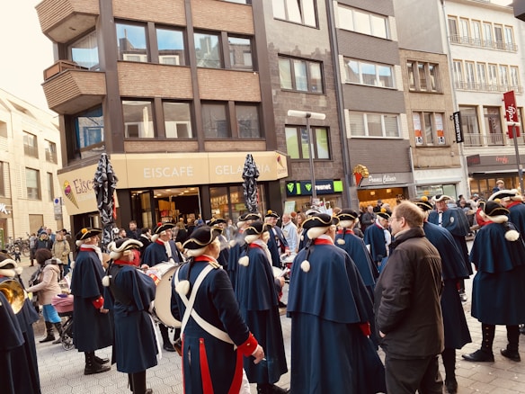 A group of people dressed in historical uniforms, featuring navy blue cloaks and tricorn hats, are gathered in a city street. They appear to be participating in a parade or public event. The background features various shops, including an Eiscafé and other businesses, set in a row of multi-story buildings.