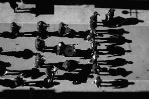 A group of musicians walking along a paved street, creating long shadows under the sunlight. Each musician appears to be playing an instrument such as a trumpet or drum, and the perspective is from above.