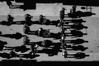 A group of musicians walking along a paved street, creating long shadows under the sunlight. Each musician appears to be playing an instrument such as a trumpet or drum, and the perspective is from above.