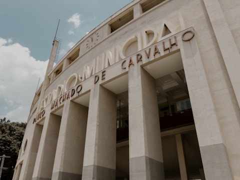 A large, imposing structure with classical architectural features such as tall, narrow columns and curving facades. The building is adorned with lettering that spells out 'Estádio Municipal Paulo Machado De Carvalho'. The image is taken from a low angle, emphasizing the verticality of the façade. The sky is partly cloudy, providing a soft backdrop to the neutral tones of the structure.