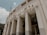 A large, imposing structure with classical architectural features such as tall, narrow columns and curving facades. The building is adorned with lettering that spells out 'Estádio Municipal Paulo Machado De Carvalho'. The image is taken from a low angle, emphasizing the verticality of the façade. The sky is partly cloudy, providing a soft backdrop to the neutral tones of the structure.