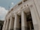 A large, imposing structure with classical architectural features such as tall, narrow columns and curving facades. The building is adorned with lettering that spells out 'Estádio Municipal Paulo Machado De Carvalho'. The image is taken from a low angle, emphasizing the verticality of the façade. The sky is partly cloudy, providing a soft backdrop to the neutral tones of the structure.