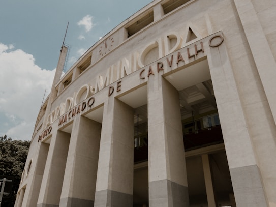A large, imposing structure with classical architectural features such as tall, narrow columns and curving facades. The building is adorned with lettering that spells out 'Est&aacute;dio Municipal Paulo Machado De Carvalho'. The image is taken from a low angle, emphasizing the verticality of the fa&ccedil;ade. The sky is partly cloudy, providing a soft backdrop to the neutral tones of the structure.