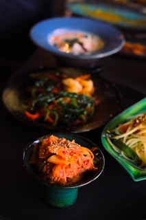 On a dark table, several dishes of Korean food are displayed. A small green bowl is filled with kimchi topped with sesame seeds. Behind it, there is a plate of saut&eacute;ed shrimp with vegetables and a blue bowl containing soup. On the right, a dish of what appears to be cold noodle salad is visible.