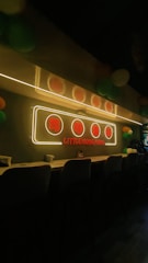A cozy interior with a long counter lined with stools. A neon sign with Chinese characters and the text 'Little Hong Kong' glows above the counter. The room is dimly lit, adding to a warm and inviting atmosphere, with colorful balloons visible in the background.