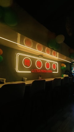 A cozy interior with a long counter lined with stools. A neon sign with Chinese characters and the text 'Little Hong Kong' glows above the counter. The room is dimly lit, adding to a warm and inviting atmosphere, with colorful balloons visible in the background.