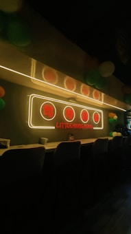 A cozy interior with a long counter lined with stools. A neon sign with Chinese characters and the text 'Little Hong Kong' glows above the counter. The room is dimly lit, adding to a warm and inviting atmosphere, with colorful balloons visible in the background.