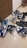 A collection of various sneakers is arranged on a tiled floor. The sneakers come in different styles and colors, featuring brands like Nike and Adidas. The shoes exhibit hues such as blue, black, white, and a pair in gray with a yellow sole.
