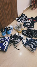 A collection of various sneakers is arranged on a tiled floor. The sneakers come in different styles and colors, featuring brands like Nike and Adidas. The shoes exhibit hues such as blue, black, white, and a pair in gray with a yellow sole.