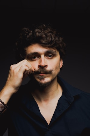 A person with curly dark hair and a carefully styled mustache is adjusting it with one hand. They are wearing a dark shirt with an open collar and a bracelet on the wrist. The background is dark, emphasizing the person's features and attire.