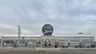A classic diner with a retro design features a prominent circular sign labeled 'I-70 Diner'. The exterior is polished metal with diamond patterns and horizontal stripes, evoking a vintage American style. Large windows line the building, and picnic benches are placed in front. The sky is overcast, with muted colors and a quiet, nostalgic atmosphere.
