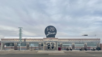A classic diner with a retro design features a prominent circular sign labeled 'I-70 Diner'. The exterior is polished metal with diamond patterns and horizontal stripes, evoking a vintage American style. Large windows line the building, and picnic benches are placed in front. The sky is overcast, with muted colors and a quiet, nostalgic atmosphere.