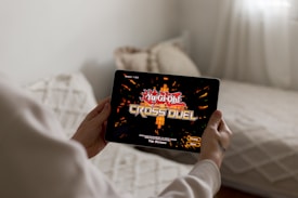 A person is holding a tablet displaying the game 'Yu-Gi-Oh! Cross Duel' on the screen. The background shows a blurred home setting with a couch and decorative cushions.