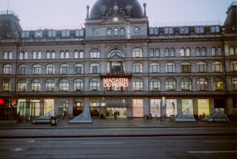 The image features a grand building with ornate architecture and a sign that reads 'Magasin Du Nord' illuminated above the entrance. The facade includes multiple arched windows across several floors. On the ground level, there are entrance doors and display windows. Small pyramid structures are positioned in front of the building along with some bicycles.