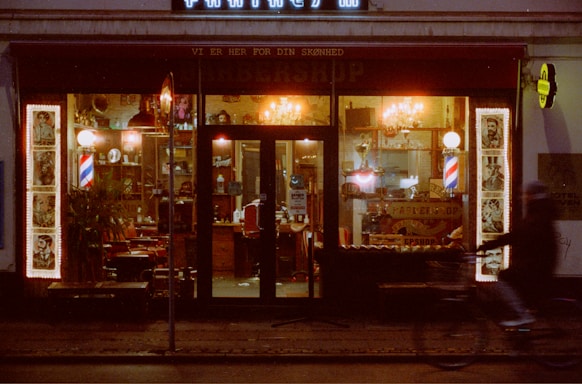 A cozy barbershop with a vintage aesthetic, featuring red and white striped barber poles, framed portraits on the outside wall, and warmly lit interior. The shop window displays classic barber chairs and a cluttered interior with various barber tools and decorations. A blurred silhouette of a person on a bicycle passes by on the street.