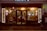 A cozy barbershop with a vintage aesthetic, featuring red and white striped barber poles, framed portraits on the outside wall, and warmly lit interior. The shop window displays classic barber chairs and a cluttered interior with various barber tools and decorations. A blurred silhouette of a person on a bicycle passes by on the street.
