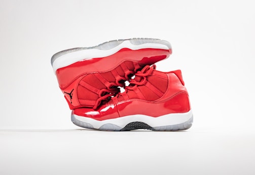 A pair of bright red high-top athletic shoes is positioned in a dynamic pose against a simple white background. The shoes feature a glossy finish with a distinct logo in black on the side. The soles are thick and white with a semi-transparent section.
