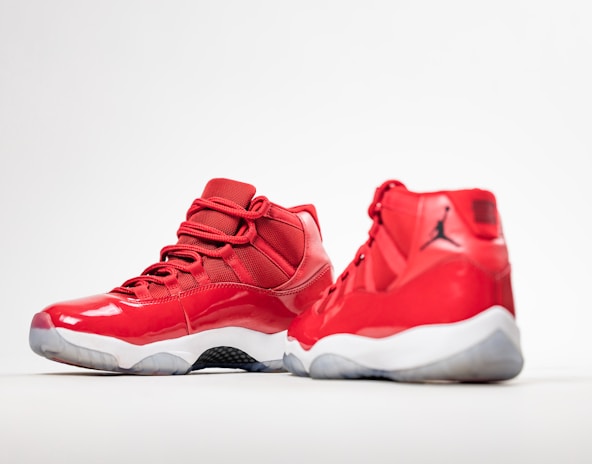 A pair of red high-top sneakers with glossy finish and white soles. One shoe is positioned facing forward, while the other is angled to show the side profile. The design features mesh fabric and a distinct black logo on the heel.