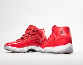 A pair of red high-top sneakers with glossy finish and white soles. One shoe is positioned facing forward, while the other is angled to show the side profile. The design features mesh fabric and a distinct black logo on the heel.