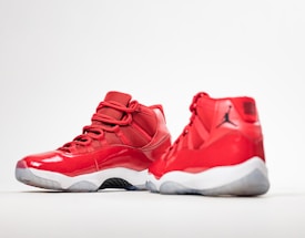 A pair of red high-top sneakers with glossy finish and white soles. One shoe is positioned facing forward, while the other is angled to show the side profile. The design features mesh fabric and a distinct black logo on the heel.