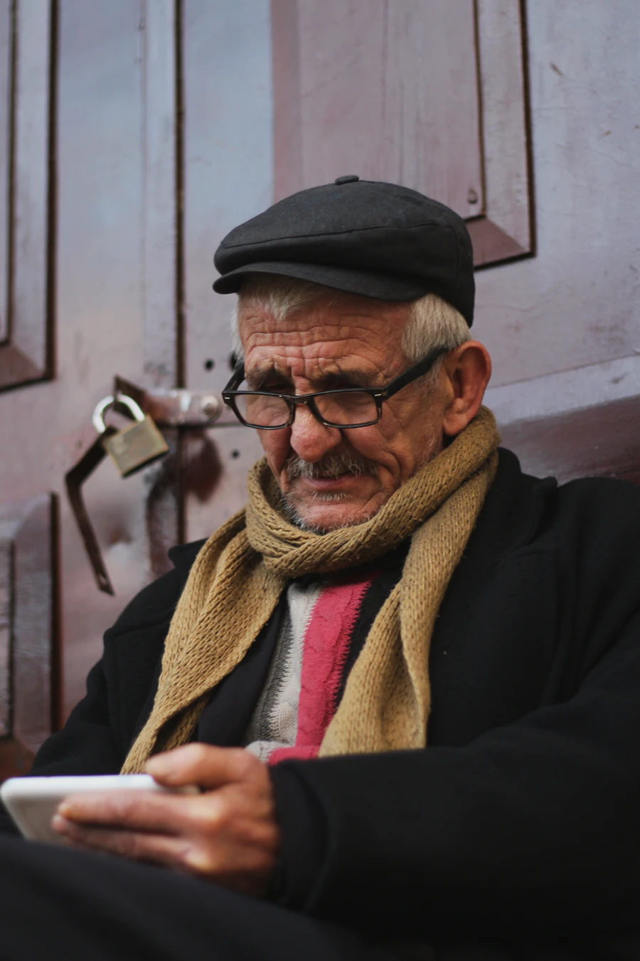 An older man in glasses and a scarf, holding a phone, looking at it with quiet attention.