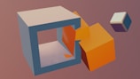 A 3D render features a large, hollow, light gray cuboid with an orange cube partially placed inside it. Another smaller cube, colored in a gradient of purple and white, floats nearby on a plain muted background.