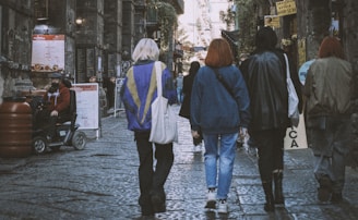 A group of people is walking down a cobblestone street lined with various shops and signs. They wear casual clothing and carry bags. One individual on the left is seated on a scooter. The setting appears urban and slightly worn with a historic charm.