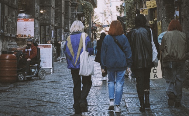 A group of people is walking down a cobblestone street lined with various shops and signs. They wear casual clothing and carry bags. One individual on the left is seated on a scooter. The setting appears urban and slightly worn with a historic charm.